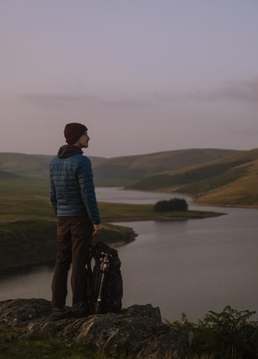 Man with a backpack on the ground next to him, stood looking out across a landscape of mountains and a lake.