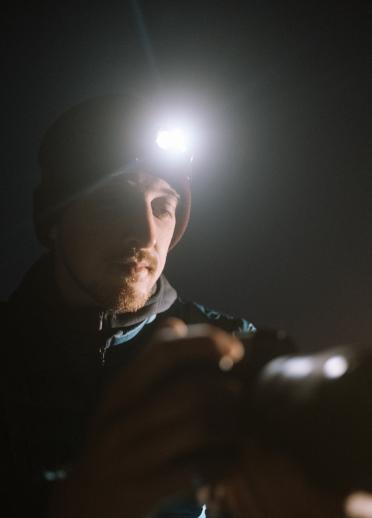 A man with a beard wearing a head torch which illuminates his face. 