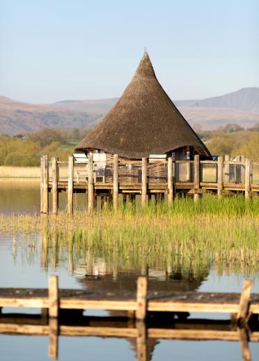 A wood-walled and reed-roofed crannog hut on a platform on a lake.