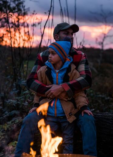 Vater und Sohn sitzen am Lagerfeuer mit Sonnenuntergang im Hintergrund.