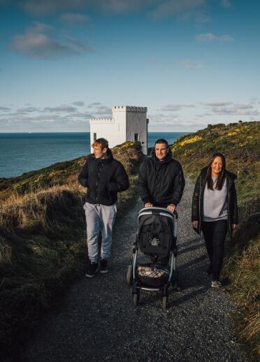 two men, one with pram, and woman walking along coast path, with white building in background. 