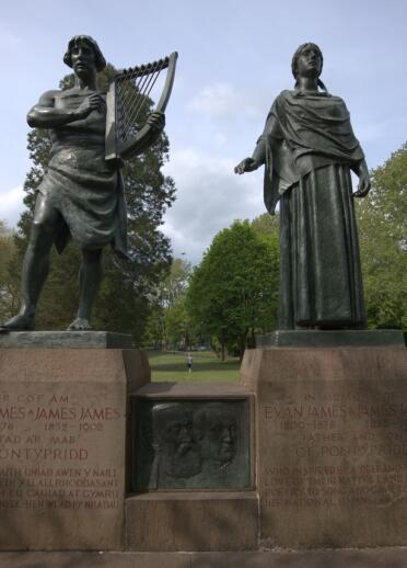 Two sculptures made of bronze. A man is holding a harp and a woman stands next to him. The statues are in a green leafy park. 