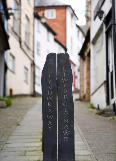 Eine stehende Schiefertafel mitten in einer schmalen Straße mit eingraviertem Text 'Glyndŵr’s Way / Llwybr Glyndŵr'.