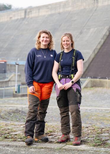 Two women in outdoor work wear stood below a dam.
