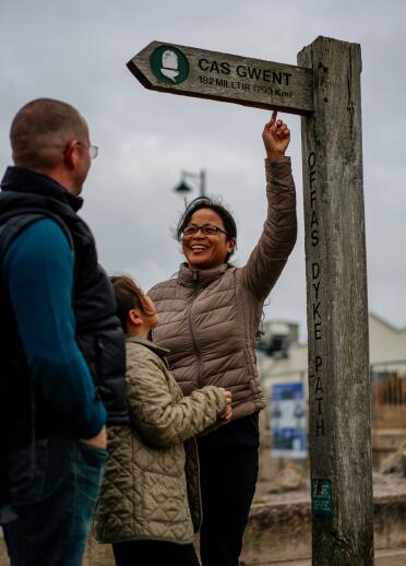 A woman pointing up at a wooden signpost with 'Casgwent' text on it.
