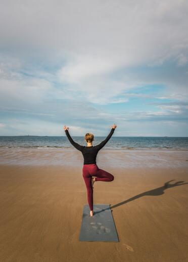 Person seen from behind, stood in a yoga pose on the beach looking out to sea.