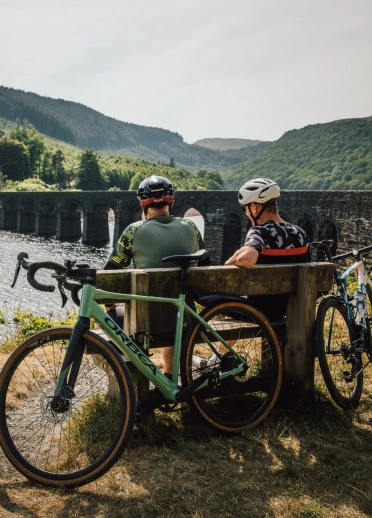Two cyclists sat on a bench by a multi-arched bridge over a lake.