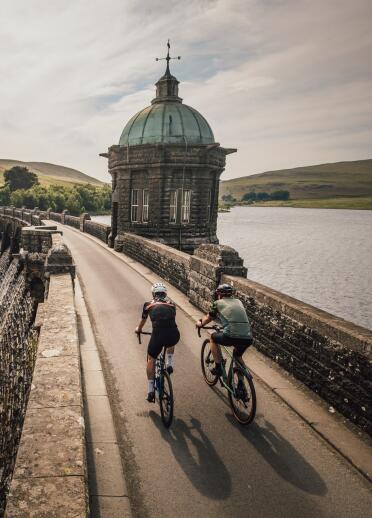 Two cyclists cycling across a dam with an ornate building on one side.