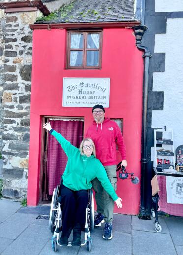 A man standing, and a woman sat in a wheelchair outside a very small red painted house.