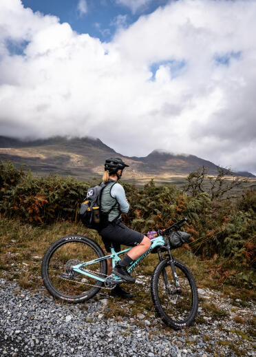 A female mountain biker on a mountainside gravel road, looking out over towards other mountains.
