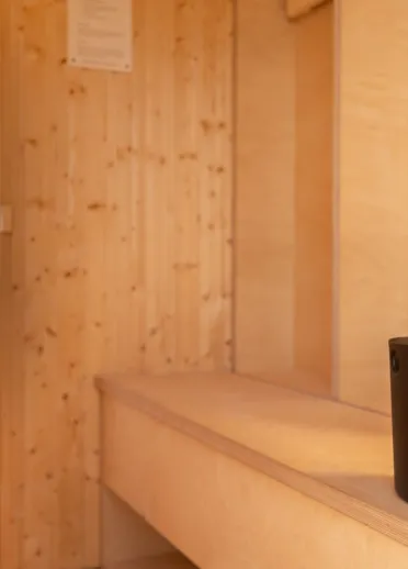 The inside of a wooden sauna with wooden walls and bench with a black bucket with wooden utensil in it.