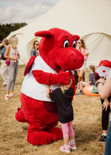 person in dragon costume hugging young child with tent and people in background.