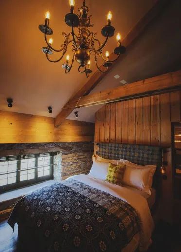 Warm rustic bedroom featuring exposed beams, wood-panelled walls, a large bed with patterned bedding, and an ornate chandelier casting soft light.