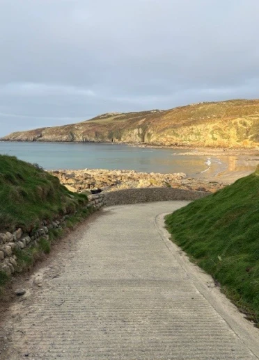 A narrow paved path leads downhill toward a calm bay, with rocky cliffs and green grassy slopes on either side. Sunlight highlights the golden cliffs in the distance under a cloudy sky.