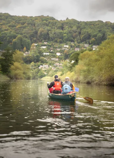 Two people paddling a canoe along a calm river, with wooded hills and scattered houses rising in the background.