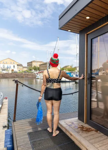 Person in swimwear standing on a marina pontoon beside a sauna cabin, holding a towel and looking out across boats and waterside buildings.