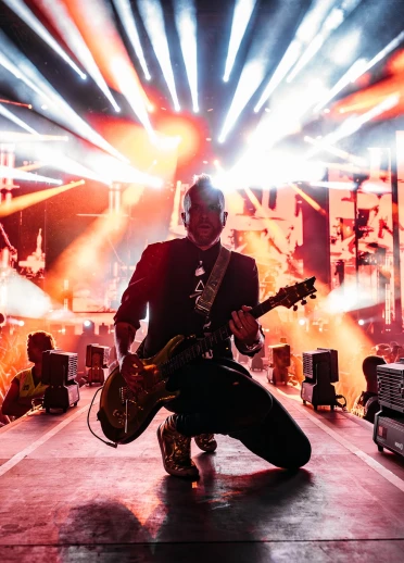Ein Mann mit Guitarre, der im Gegenlicht auf einer Bühne kniet. Im Hintergrund viele Lichter und Personen.