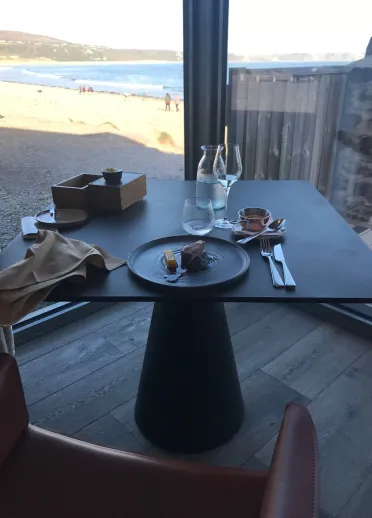 A round black table and chairs sit in the foreground, while outdoor decking and coastal views stretch beyond the glass under a bright sky.