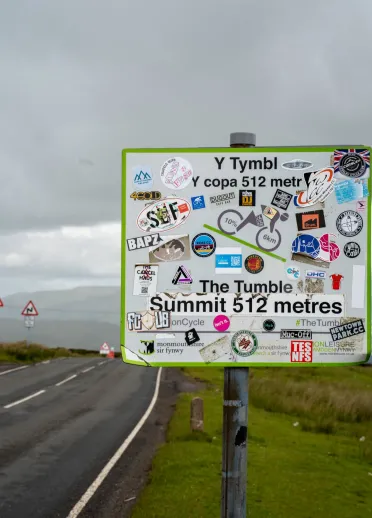 A heavily stickered road sign with 'Y Tymbl y copa / The Tumble summit' text.