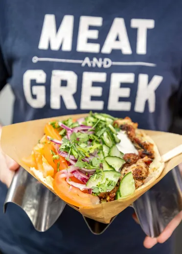 Person wearing a “MEAT AND GREEK” T-shirt holding a Greek-style flatbread filled with grilled meat and salad.