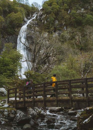 People walking over a footbridge over a stream looking up at a waterfall.