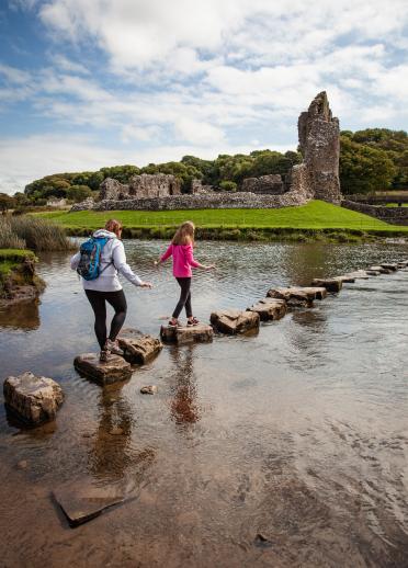 two young females walking on stepping stones with castle ruins in the background.