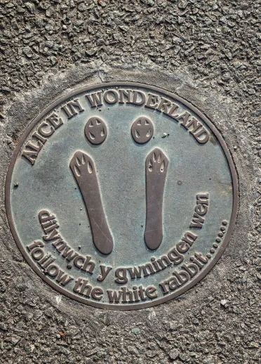 An Alice Town Trail metal sign inset into the pavement - rabbit footprints with 'follow the white rabbit' text.