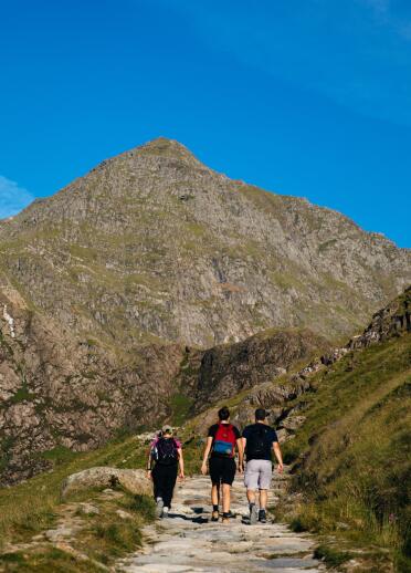 Drei Wanderer auf einem gepflasterten Weg, der auf einen hohen Gipfel führt.