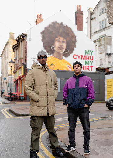 Two men in front of a mural on the side of a building. The mural shows a Black lady wearing a yellow Wales football shirt.
