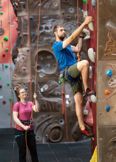 A man climbing up an indoor climbing wall with a woman holding the rope.