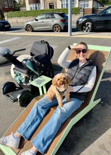 woman laid out on a fixed sun lounger-style chair on promenade, shielding her face from the sun with her wheelchair next to her and dog on her knee.