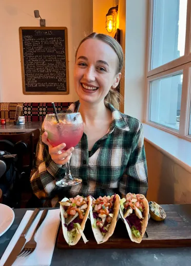 woman sat at table with three sea bream tacos on the plate in front of her and holding up a pink mocktail. 