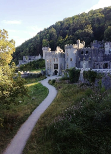A stone castle in a green valley, with a winding footpath leading toward the building and wooded hills rising behind it.