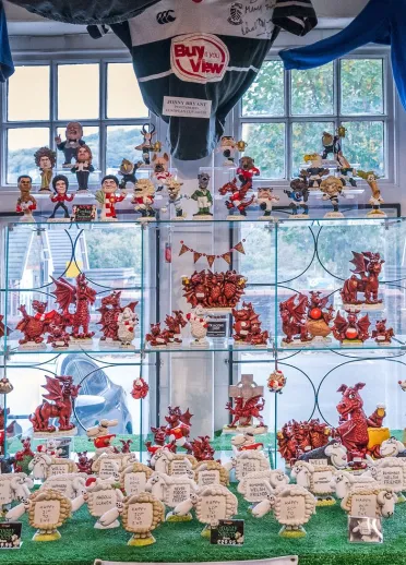 A shop display filled with Welsh dragon figurines and sheep souvenirs arranged on glass shelves beneath hanging rugby shirts.