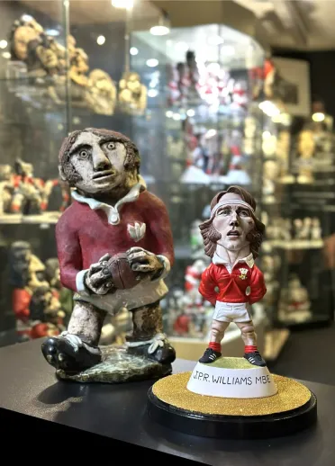 Two rugby‑themed ceramic figurines displayed in a glass‑filled showroom, one holding a rugby ball and the other standing on a base labelled “J.P.R. WILLIAMS MBE”.