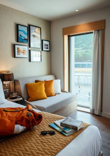 A hotel room with adventure books and rucksack on the bed and views of surfers on a lagoon.