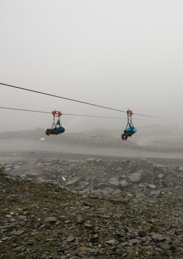 Four people attached with safety harnesses riding a zip line over a slate quarry, with a misty sky line.