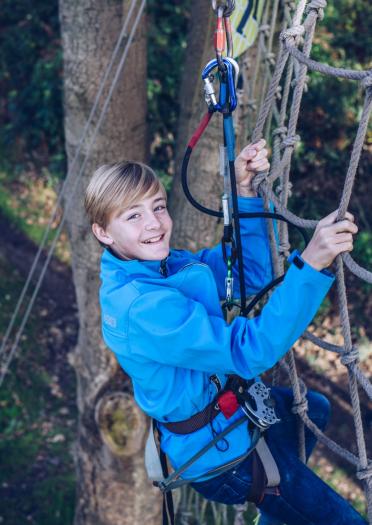 Junge mit Klettergeschirr klettert über ein Netz hoch in einem Baum.