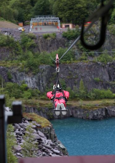 A person flying down on Zip World Velocity zip wire with the blue waters of Penrhyn Quarry beneath.