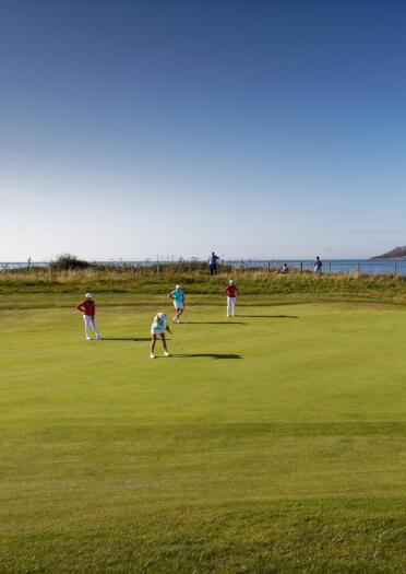 Golfers putting on a green with views of the coastline beyond.