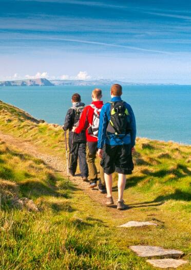 Drei Männer in Wanderausrüstung wandern entlang des Wales Coast Path mit Blick auf das Meer.