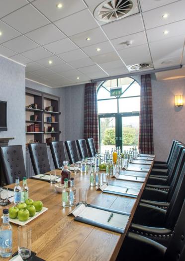 A long boardroom table with fruit, water and stationery surrounded by high back chairs.