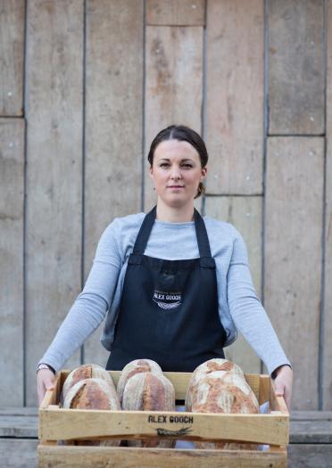 Woman holding box of freshly baked loaves looks straight at the camera.