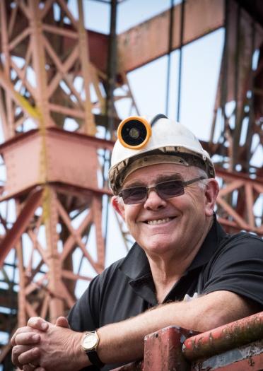 A former miner smiling at the camera with a tower colliery behind him.