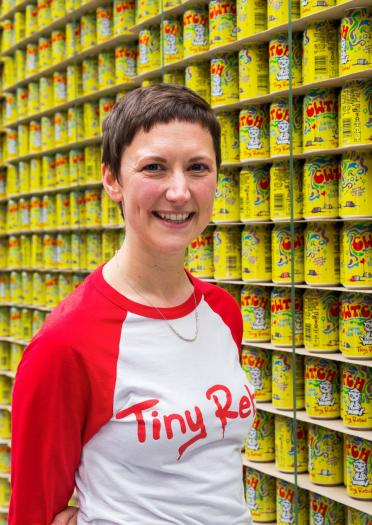 female in branded tshirt stood infront of stacked shelf full with yellow Tiny Rebel cans.