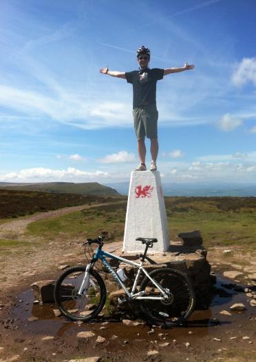 Man standing on top of the Hay Bluff trig point with a bike underneath.