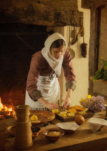 woman dressed in 17th century period costume attending to table of food with fire in the background