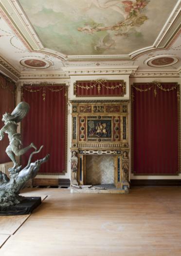 Drawing room inside Dyffryn House with red walls, ornate ceiling and a statue in the foreground.