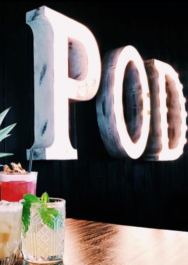 Cocktails und Obst im Vordergrund mit beleuchtetem Schild "POD" im Hintergrund.