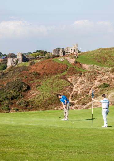 Golfer, der einen Putt schlägt, mit einem Golfer, der die Flagge auf dem Platz des Pennard Golf Clubs hält, mit Pennard Castle im Hintergrund.
